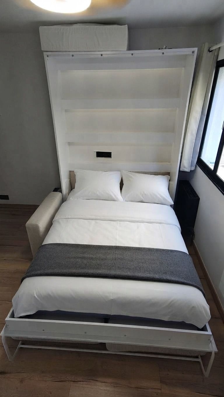 Le lit escamotable 140 × 200 déplié et fait : linge blanc, plaid gris, étagères en tête de lit, accoudoir du canapé à gauche et fenêtre à menuiserie noire à droite.