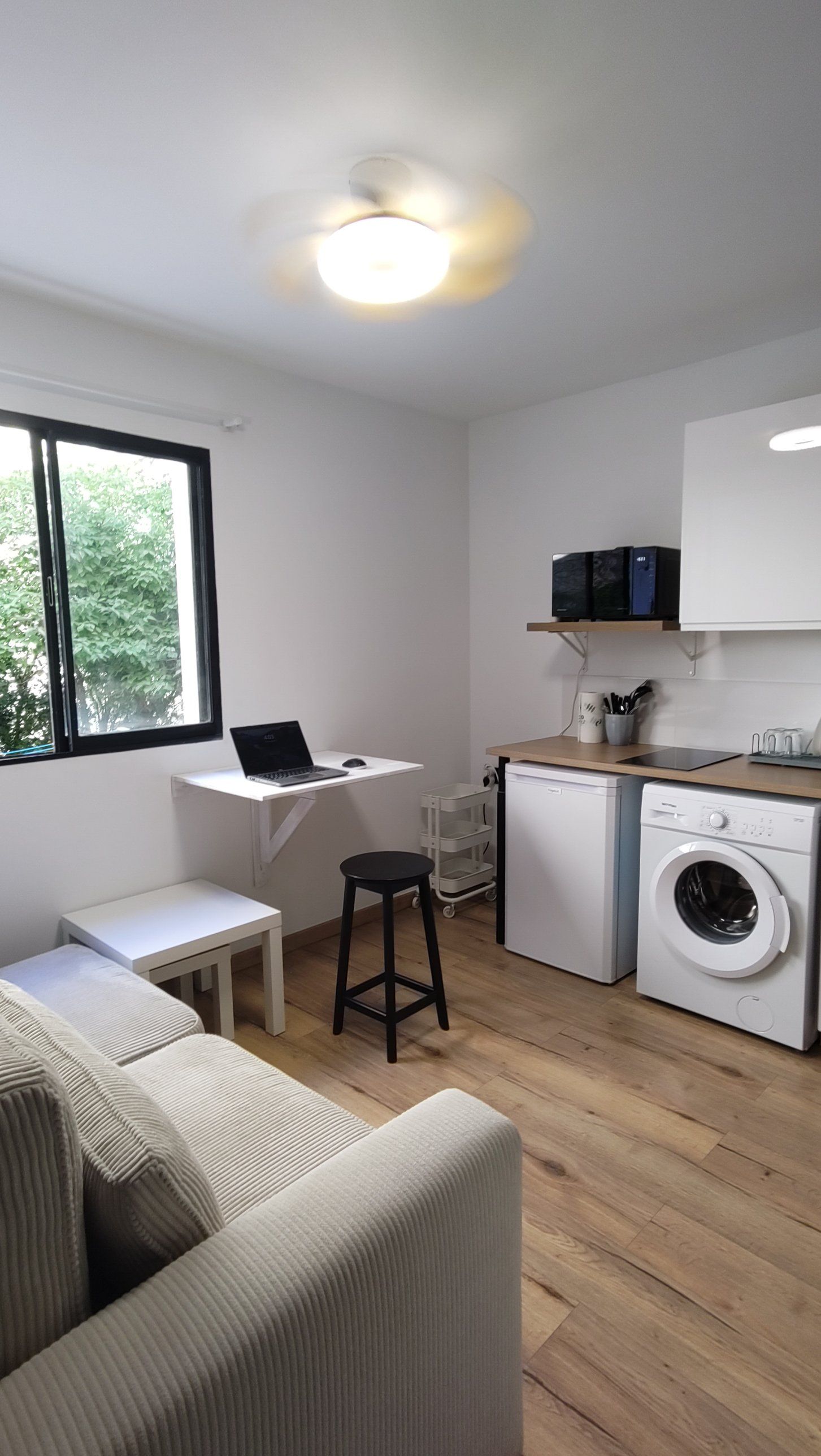 Le séjour : une table murale rabattable avec un ordinateur portable, un tabouret, la kitchenette et le lave-linge, sous la fenêtre.