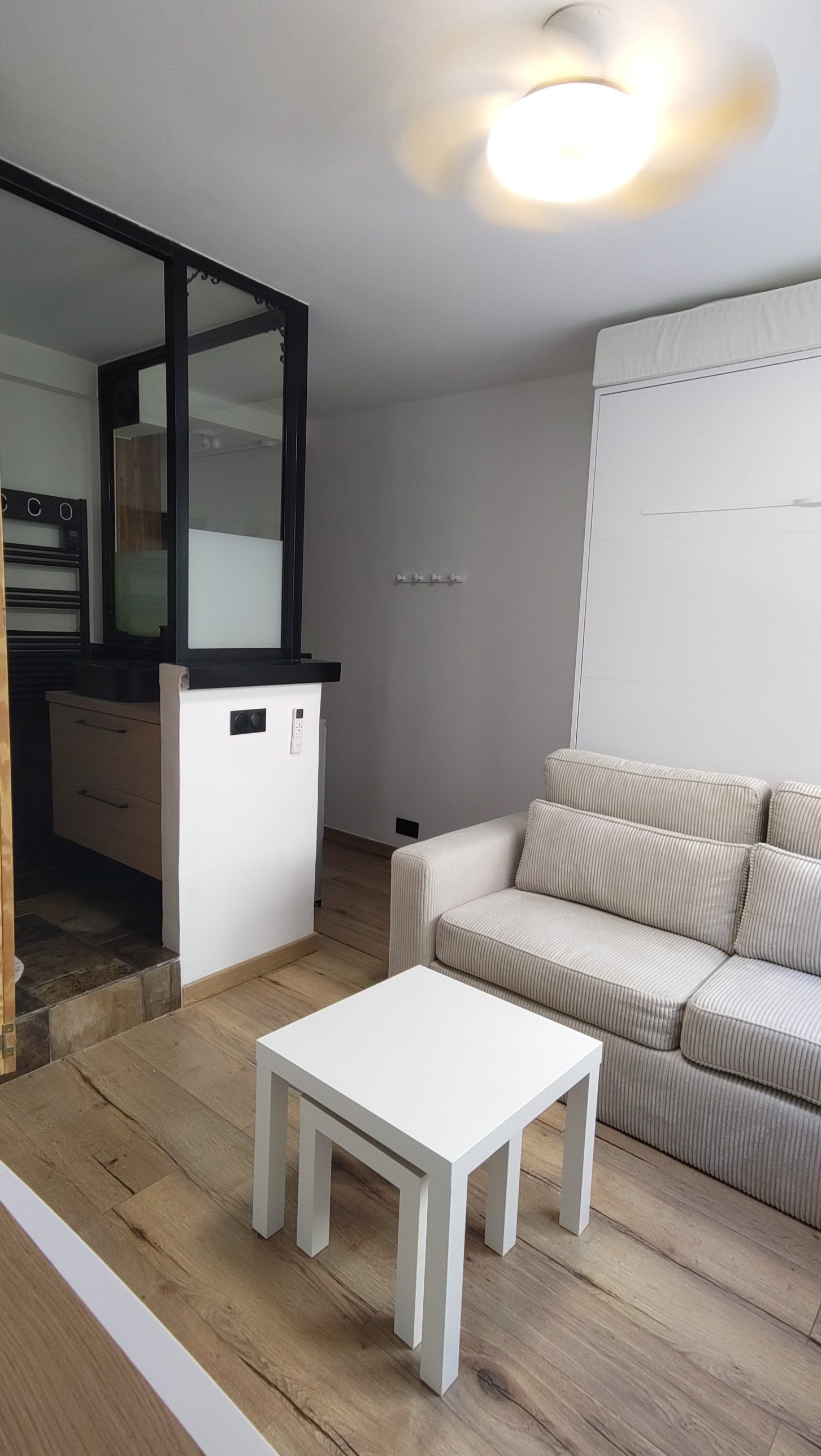 Le studio en configuration jour : lit replié dans son meuble, canapé et table basse, salle de bain derrière la verrière noire.